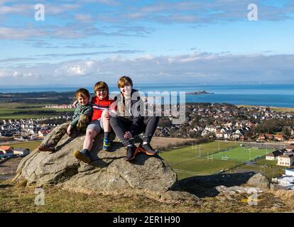 North Berwick, East Lothian, Schottland, Großbritannien, 28th. Februar 2021. Berwick-Gesetz für Rett-Syndrom: Am Tag der Seltenen Erkrankungen steigt die 7-jährige Nina McKinney diesen Monat jeden Tag auf den 613ft-Hügel, um Geld für Reverse Rett zu sammeln, eine Wohltätigkeitsorganisation, die die Erforschung der Krankheit unterstützt, an der ihre Schwester leidet. Nina hat dem Wetter und zum Glück heute der Sonne trotzt. Im Bild: Nina mit ihren beiden Brüdern Angus, 11 Jahre und Felix, 4 Jahre, sitzen auf einem Felsen mit Blick auf North Berwick Stockfoto