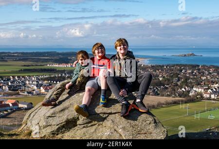 North Berwick, East Lothian, Schottland, Großbritannien, 28th. Februar 2021. Berwick-Gesetz für Rett-Syndrom: Am Tag der Seltenen Erkrankungen steigt die 7-jährige Nina McKinney diesen Monat jeden Tag auf den 613ft-Hügel, um Geld für Reverse Rett zu sammeln, eine Wohltätigkeitsorganisation, die die Erforschung der Krankheit unterstützt, an der ihre Schwester leidet. Nina hat dem Wetter und zum Glück heute der Sonne trotzt. Im Bild: Nina mit ihren beiden Brüdern Angus, 11 Jahre und Felix, 4 Jahre, sitzen auf einem Felsen mit Blick auf North Berwick und halten für eine Pause, während sie schauen, was noch zu klettern übrig ist Stockfoto