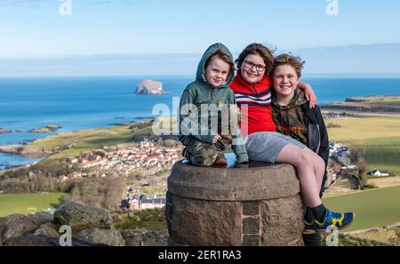 North Berwick, East Lothian, Schottland, Großbritannien, 28th. Februar 2021. Berwick-Gesetz für das Rett-Syndrom: Die siebenjährige Nina McKinney beendet jeden Tag in diesem Monat ihr Ziel, den 613ft-Hügel zu erklimmen, der Rest eines vulkanischen Stopfens, um Geld für Reverse Rett zu sammeln, eine Wohltätigkeitsorganisation, die die Erforschung der Krankheit unterstützt, unter der ihre Schwester leidet. Im Bild: Nina mit ihren beiden Brüdern Angus, 11 Jahre und Felix, 4 Jahre, erreichen die Spitze des Hügels, um die Spendenaktion mit Blick auf den Bass Rock im Firth of Forth im Hintergrund abzuschließen Stockfoto