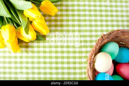 Gelbe Tulpen und Korb mit bunten Eiern über schäbig karierter grüner Tischdecke. Draufsicht. Speicherplatz kopieren. Stockfoto