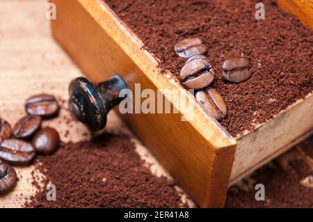 Geöffnete Schublade einer manuellen Kaffeemühle aus Holz mit gemahlenem Kaffeepulver im Inneren und einzelnen gerösteten Kaffeebohnen mit geringer Schärfentiefe. Stockfoto