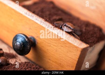 Geöffnete Schublade einer manuellen Kaffeemühle aus Holz mit gemahlenem Kaffeepulver im Inneren und einzelnen gerösteten Kaffeebohnen mit geringer Schärfentiefe. Stockfoto