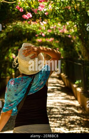 Eine junge kaukasische Frau, die einen großen Eimerhut trägt Aus locker gestricktem Raffia ist allein in einem zu Fuß Gartenpfad im Alhambra Palast mit schöner Stockfoto