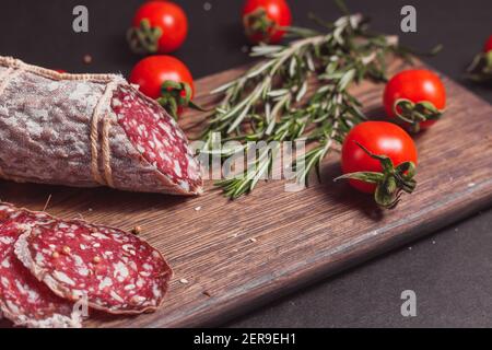 Geräucherte Schinkenwurst und geschnittene Scheiben mit Rosmarin und Kirsche Tomaten auf dunklem Holzbrett Stockfoto