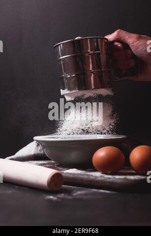 Hand hält Metallmehlsieb, ein Sieb bestreut Mehl. Mehl sieben. Vorbereitung zum Backen. Low-Key-Foto Stockfoto