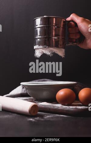 Hand hält Metallmehlsieb, ein Sieb bestreut Mehl. Mehl sieben. Vorbereitung zum Backen. Low-Key-Foto Stockfoto