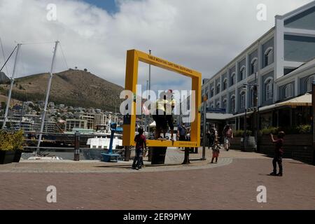 KAPSTADT, Südafrika 30. Januar 2021: Kleiner Junge, der ein Foto mit einem Handy seiner Eltern in einem großen gelben Rahmen am Ufer von V&A machte. Stockfoto