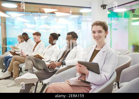 Porträt einer jungen Frau im Labormantel und lächelnd an der Kamera, während sie in Reihe mit einer multiethnischen Gruppe von Menschen im Publikum bei einem medizinischen Seminar sitzt, Stockfoto