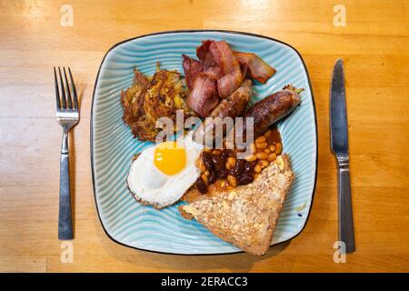 Ein traditionelles englisches Frühstück auf einem blauen Teller mit Würstchen, einem Spiegelei, Schinkenspeck, einem Rösti, Bohnen und Toastscheiben Stockfoto