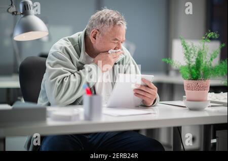 Während der Coronavirus-Pandemie sitzt ein erwachsener Mann in einem Büro an einem Schreibtisch, liest Dokumente und hat eine laufende Nase Stockfoto