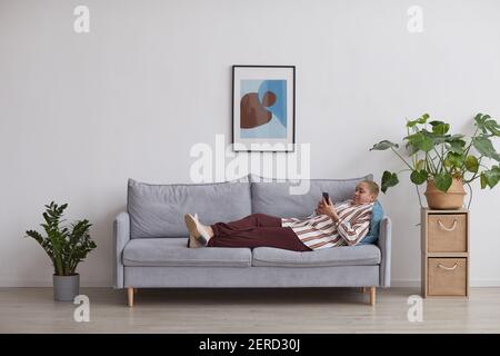 Weitwinkel-Ansicht auf moderne Mixed-Race-Frau mit Smartphone, während auf dem Sofa in minimalem Zuhause Interieur liegen, kopieren Platz Stockfoto