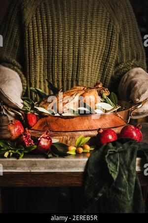 Frau in Strickpullover und Küchenhandschuhe halten ganze gebratene Huhn für den Urlaub festliches Abendessen in Kupfer-Bräter mit Gewürzen, Kräutern und Fr. Stockfoto