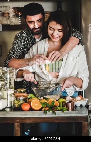 Junge glückliche Paar Kochen zusammen in der Küche. Kaukasischen Mann Gesicht in Mehl und Brünette Frau Umarmung, Flirten, Lachen und Mischen Zutaten für Stockfoto