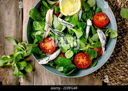 Grüner Feldsalat mit eingelegten Sardellen oder Sardinenfilet und Kirschtomaten, serviert in blauer Schüssel mit Zitrone und Olivenöl auf Strohserviette über ol Stockfoto