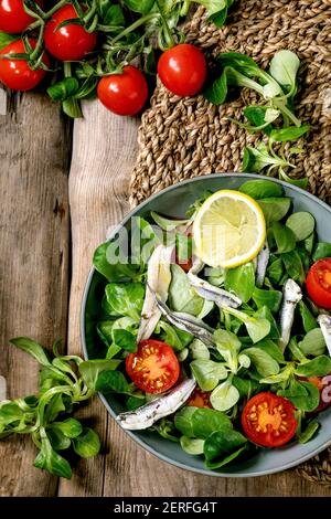 Grüner Feldsalat mit eingelegten Sardellen oder Sardinenfilet und Kirschtomaten, serviert in blauer Schüssel mit Zitrone und Olivenöl auf Strohserviette über ol Stockfoto