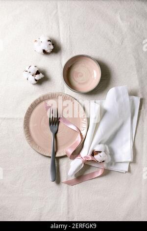 Set von leeren rosafarbenen Keramikplatten mit Gabel und Textilserviette, verziert mit Baumwollblumen und Seidenband über weißer Leinentischdecke. Tabelleneinstellung. Stockfoto