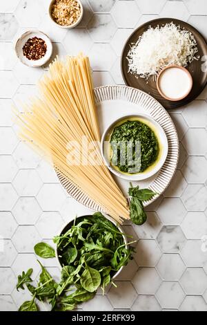 Linguini auf einem Teller mit einer Schüssel Basilikumpesto trocknen, neben einer Schüssel frischen Spinat, geriebenen Parmesankäse, zerkleinerten roten Pfeffer und gerösteten Brotkrumen Stockfoto