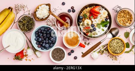 Gesundes Frühstücksset. Hafergranola mit frischen Beeren, Banane, Joghurt, Ahornsirup, Samen und Minzblättern mit Tasse Kaffee auf rosa Hintergrund, flach Stockfoto