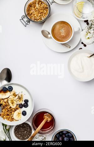 Gesundes Frühstücksset. Hafergranola mit Joghurt, Honig, frischen Bananen, Heidelbeeren, Chiasamen in Schüssel und Tasse Kaffee auf weißem Hintergrund, Draufsicht, Stockfoto