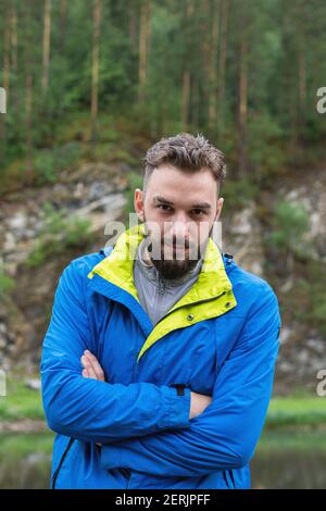Junger bärtiger Kerl im Regenmantel schaut mit seiner Kamera an Hände gefaltet auf seiner Brust im Naturpark Stockfoto