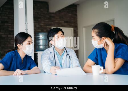 Gruppe von männlichen und weiblichen asiatischen Ärzten in Uniform trinken Kaffee trägt Gesichtsmaske und Stethoskop während der Diskussion Patientenfall Sitzen in der Kantine Stockfoto