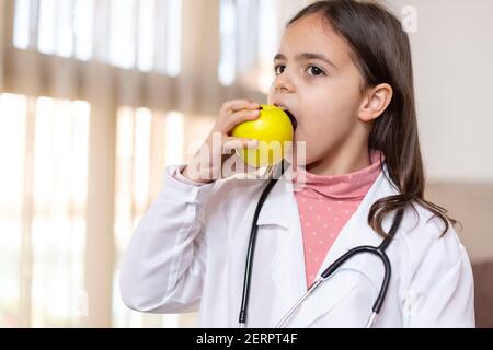 Portrait eines kleinen Mädchens, das als Arzt gekleidet ist und einen Apfel isst. Konzept von Wellness und gesundes Leben. Leerzeichen für Text. Stockfoto