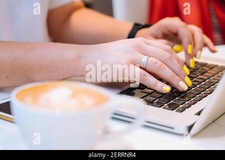 Nahaufnahme Tasse Cappuccino Kaffee und Hände Frau sitzt im Büro Coworking und tippen auf ihrem Laptop Tastatur Computer. Schreibtisch mit Telefon, Notebook, Brille und einer Tasse Kaffee. Geschäftskonzept Stockfoto