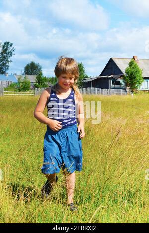 Ein Junge in blauen Shorts und einem gestreiften T-Shirt geht durch die Wiese. Dorfhäuser auf der Rückseite. Das Kind lächelt. Gute Laune. Unbeschwerte Kindheit. Stockfoto