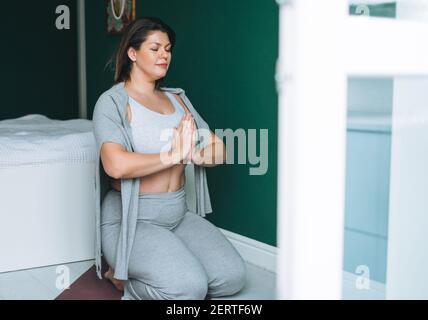 Schöne Brünette junge Frau plus Größe Körper positiv in komfortabel Sportbekleidung Yoga online zu Hause üben Stockfoto