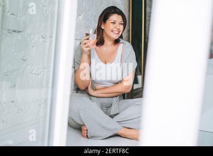 Schöne glücklich Brünette junge Frau plus Größe Körper positiv in Bequeme Sportbekleidung mit einem Glas Wasser zu Hause Stockfoto