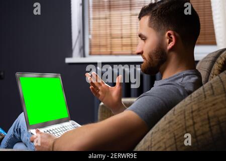 Mann sitzt entspannt auf einer Couch arbeitet auf einem Laptop mit leerem grünen Bildschirm. Dayout in seinem Wohnzimmer Mann verwendet Notebook-Computer. Office Online-Arbeit Stockfoto