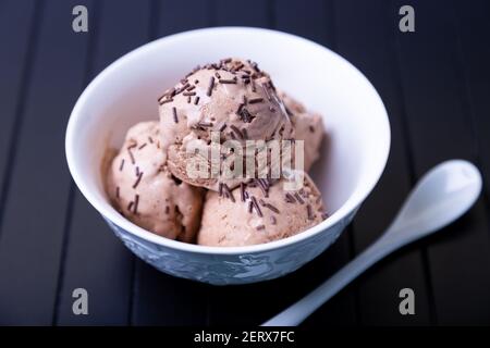 Schokoladeneis mit Schokolade in eine weiße Schüssel auf schwarzem Hintergrund streuen. Drei Kugeln hausgemachtes Eis. Nahaufnahme. Stockfoto