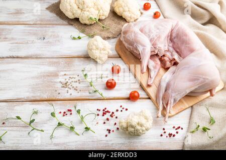 Ganz rohes Kaninchen mit Blumenkohl, Tomaten und Gewürzen auf weißem Holzhintergrund und Leinentextilien. Seitenansicht, Kopierbereich. Stockfoto