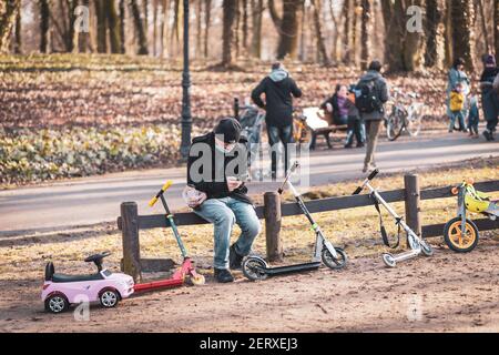 POZNAN, POLEN - 21. Feb 2021: Mann mit Gesichtsmaske und Smartphone, während er auf einem Holzzaun mit sich lehnenden Rollern auf einem Spielplatz sitzt Stockfoto