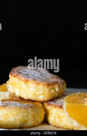 Süße Quark Käsekuchen mit orangen Scheiben auf einem weißen Teller Nahaufnahme an einer schwarzen Wand mit Kopierraum. Vertikales Foto Stockfoto