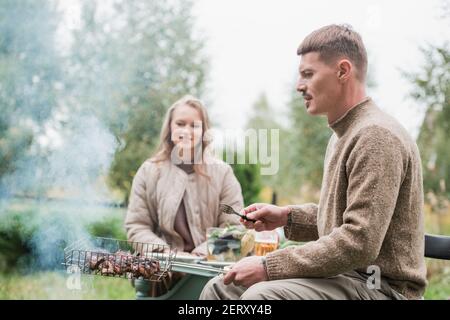 Ein Mann und eine Frau kochen gemeinsam in der Natur einen Grill. In einem Datscha-Dorf auf eigenem Grundstück. Warmes Herbstwetter, gemütliches Familienessen. Stockfoto