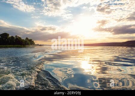 Szenische helle lebendige blau bis rot warmen Sonnenuntergang Abendzeit Landschaft mit Motorboot swirl Spur auf Wasseroberfläche des Sees oder Fluss. Horizont-Küstenlinie Stockfoto