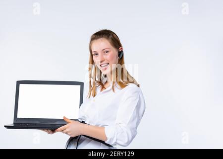 Junge Blondine mit Ohrhörer im Ohr. Zeigt der Kamera einen weißen Laptop-Bildschirm an. Weißer Hintergrund. Stockfoto