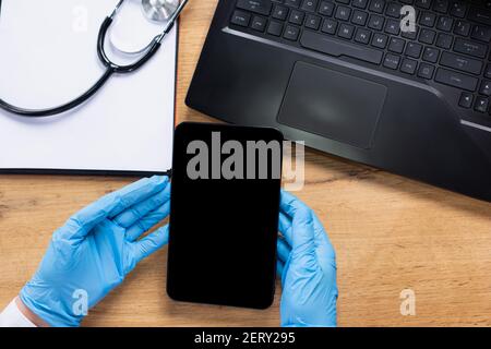 Hände eines Sanitäters in Latexhandschuhen, die eine Tablette halten. Auf dem Tisch ein Laptop, Papier und ein Stethoskop. Zugeschnittenes Foto. Stockfoto