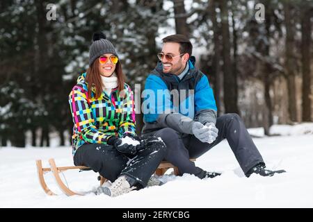 Lächelndes glückliches Paar, das auf Schlitten sitzt und Schnee in den Händen hält, Winterzeitkonzept Stockfoto
