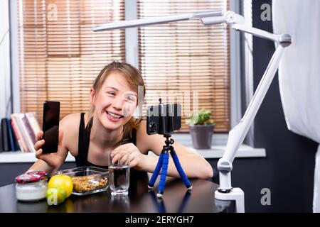 Hübsche kaukasische Blondine in der Küchentheke, Blick auf das Telefon mit einem anderen Telefon in der Hand nach dem Rezept für das Abendessen. Lächelnde Frau Stockfoto