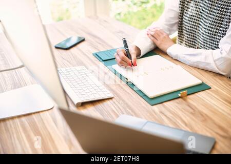 High-Angle-Aufnahme einer nicht erkennbaren Geschäftsfrau, die am Schreibtisch sitzt und etwas in ihr Notebook schreibt. Stockfoto