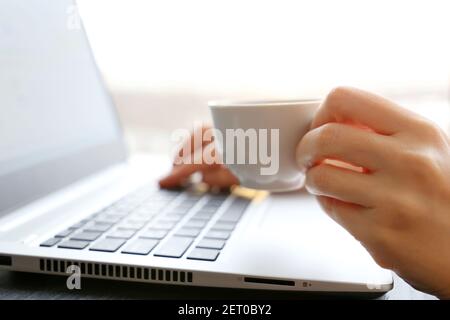 Frau trinkt Kaffee, sitzt auf Laptop-Tastatur auf Fensterhintergrund. Weiße Tasse heißes Getränk in weiblicher Hand in Sonnenschein, guten Morgen Stockfoto