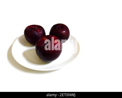 Drei süße reife saftige rote Pflaumen (prunus) auf einem Teller auf weißem Grund. Frisch gegessen, verwendet, um Marmelade zu machen oder zu Pflaumen getrocknet. Hoher Winkel. Copyspace Stockfoto