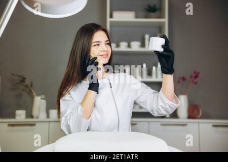 Beratung in der Kosmetologie Klinik. Weibliche Schönheitsärztin in weißem Mantel und schwarzen Handschuhen spricht am Telefon und beobachten Flasche Sahne. Gesundheitskonzept. Stockfoto