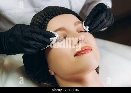 Glückliche junge schöne Frau lächelt freudig, während professionelle Kosmetikerin Reinigung ihr Gesicht mit Wattepads Vorbereitung für Gesichtsbehandlungen. Klinisch Stockfoto