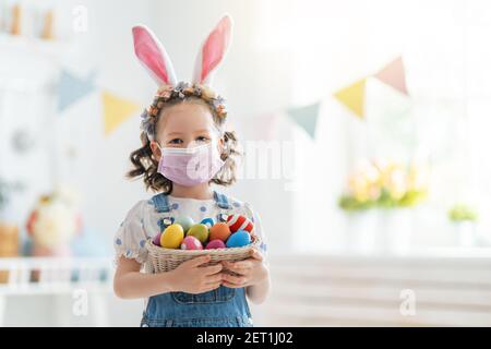 Schönes Kind mit malen Eier. Glückliche Familie bereitet sich auf Ostern vor. Nettes kleines Mädchen trägt Hasenohren und Gesichtsmaske. Stockfoto