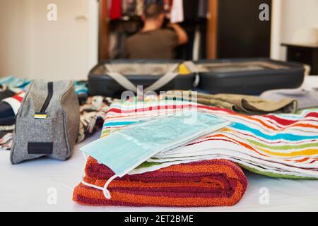Ein offener Koffer auf dem Bett, verpackt mit verschiedenen Kleidern, wie Badehosen, Strandhandtüchern und einigen blauen Einweg-Gesichtsmasken, und einem jungen Caucas Stockfoto