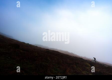 Mountainbiker am Fuße eines steilen Hügels, der in einer Wolkeninversion mit blauem Himmel über dem Berg bergauf radelt, Ilkley Moor, West Yorkshire, England, UK Stockfoto