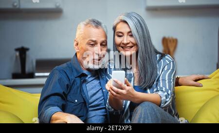 Fröhliche asiatische Frau mit Handy in der Nähe erstaunt älteren Mann Stockfoto
